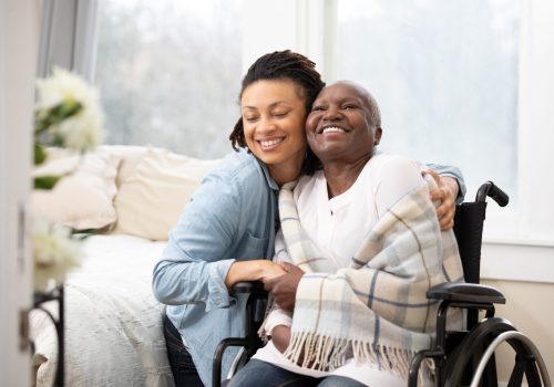 A daughter puts her arms around a smiling mother wrapped up in a blanket, sitting in a wheelchair.