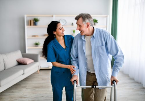 Female caregiver assisting elderly man to walk with frame at retirement home. Aged male patient with nurse moving around at seniors centre. Professional health care service for older people