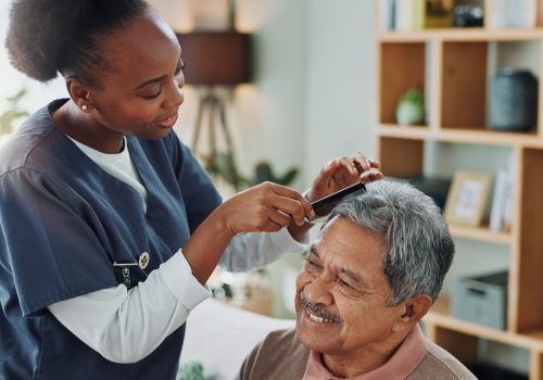 Living room, elderly man and nurse with comb for hair care, grooming or support in nursing home. Senior person, medical professional and discussion with helping for wellness, hairstyle and retirement